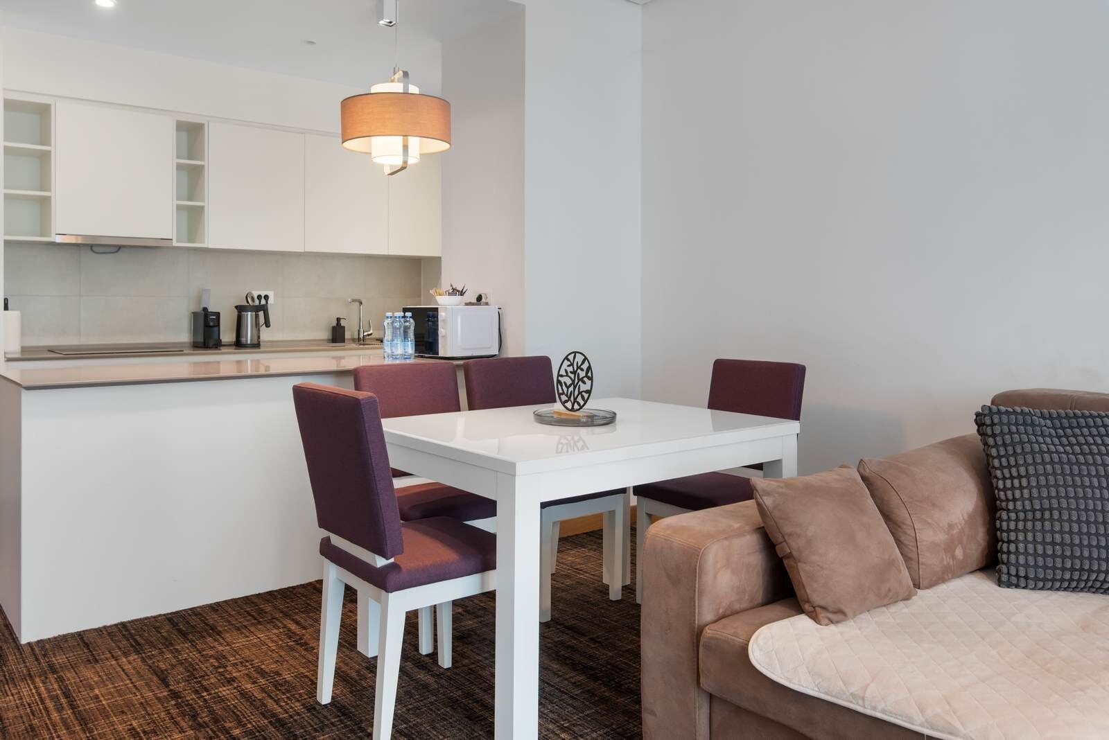 A modern kitchen with white cabinets, a dining table with chairs, a sofa, and a pendant light.