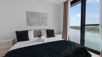 Un dormitorio de hotel con una cama grande, una mesita de noche, un ventanal con vista al río y un cuadro abstracto colgado en la pared.