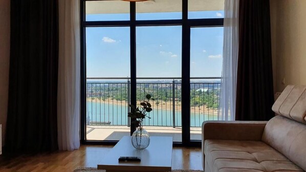 A living room with a large window, a sofa, a coffee table with a vase, and a remote control. - Belgrade Waterfront Riverside Apartments (Belgrade)