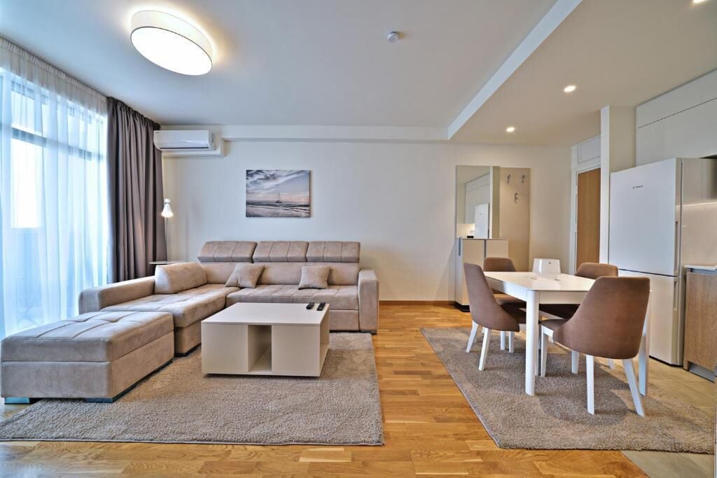 A modern living room with a sectional sofa, a coffee table, a dining area with chairs, and a kitchen with a refrigerator.