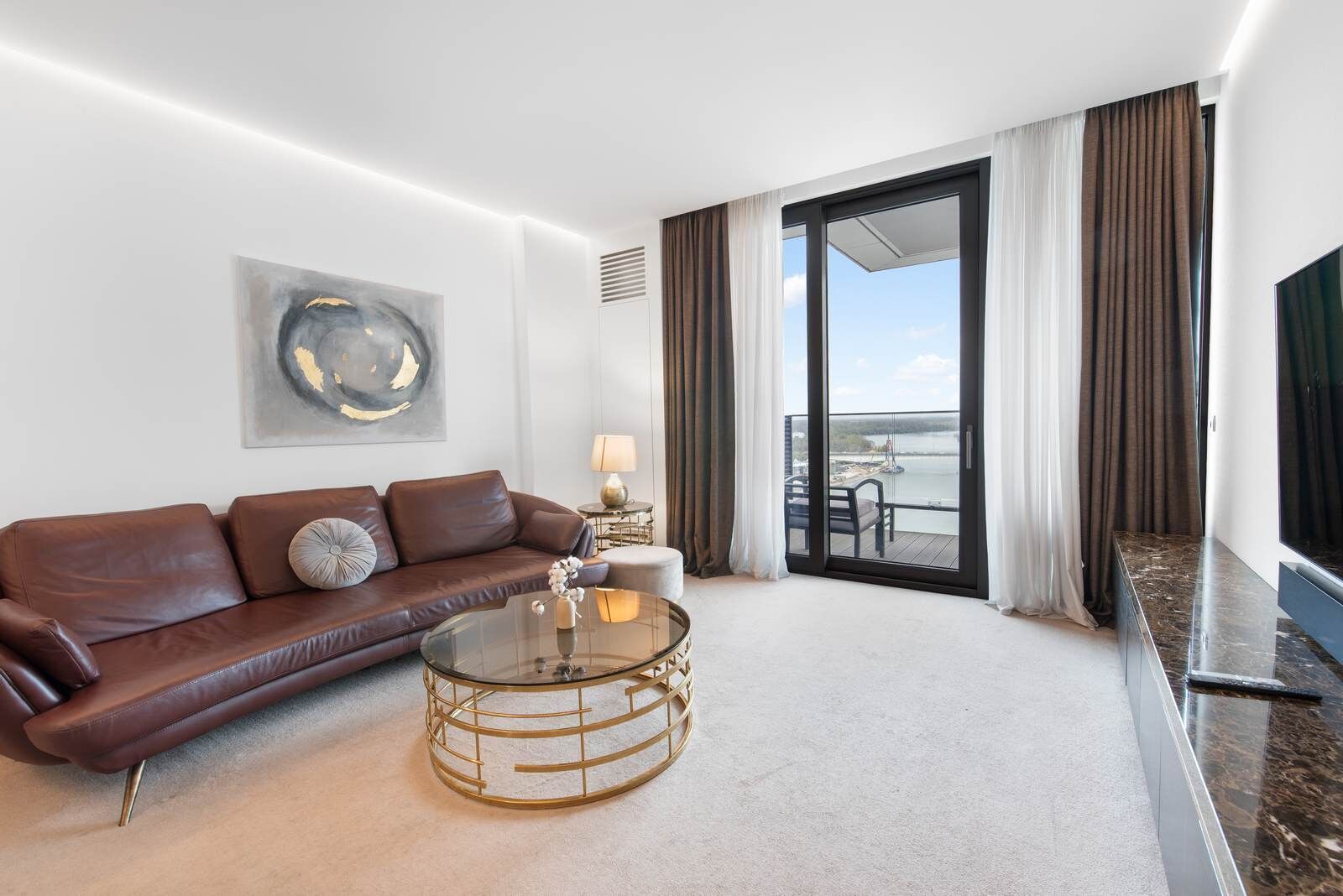 A modern hotel room with a leather sofa, a glass coffee table, a flat-screen TV, and a balcony with a view.
