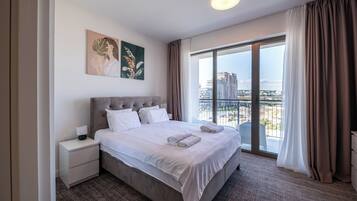 A modern hotel room with a large bed, a nightstand, a painting on the wall, and a view of the city through a large window.