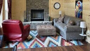 A cozy living room with a stone fireplace, wooden walls, a red armchair, a grey sofa, and a colorful rug. - 4K River Ranch • River Cabin 3 (sleeps 4) (Red River)