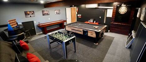A room with a foosball table, pool table, dartboard, and a dartboard.