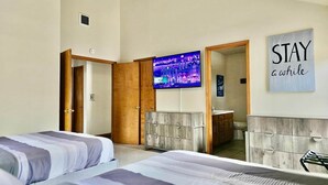 A hotel room with two beds, a TV, a ceiling fan, a mirror, and a 'STAY a while' sign.