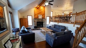 A spacious living room with a stone fireplace, wooden ceiling beams, a blue sofa set, and a staircase with wooden railings.