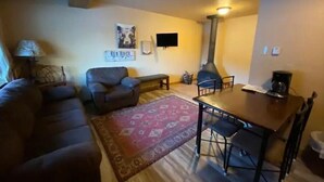 A cozy living room with a sofa, a coffee table, a dining table with chairs, a red patterned rug, a fireplace, and a wall-mounted TV. - 4K River Ranch • Condo 3 (sleeps 4) (Red River)