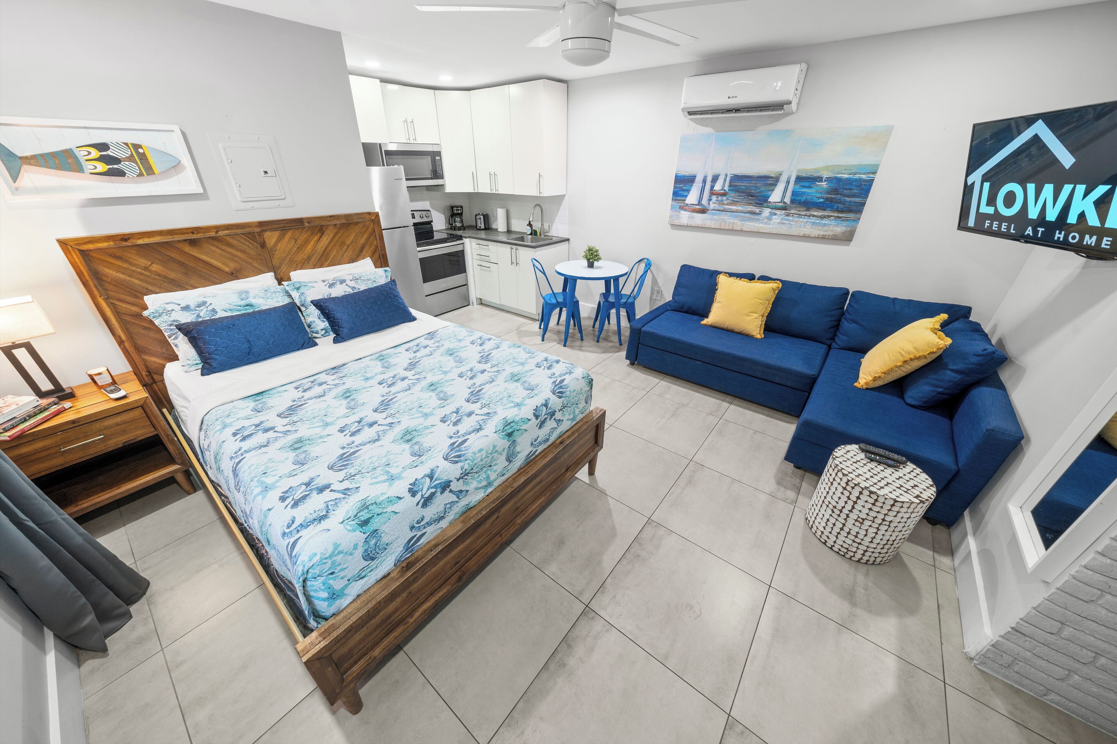 A modern hotel room with a wooden bed, a blue sofa, a small dining table, and a kitchen area.
