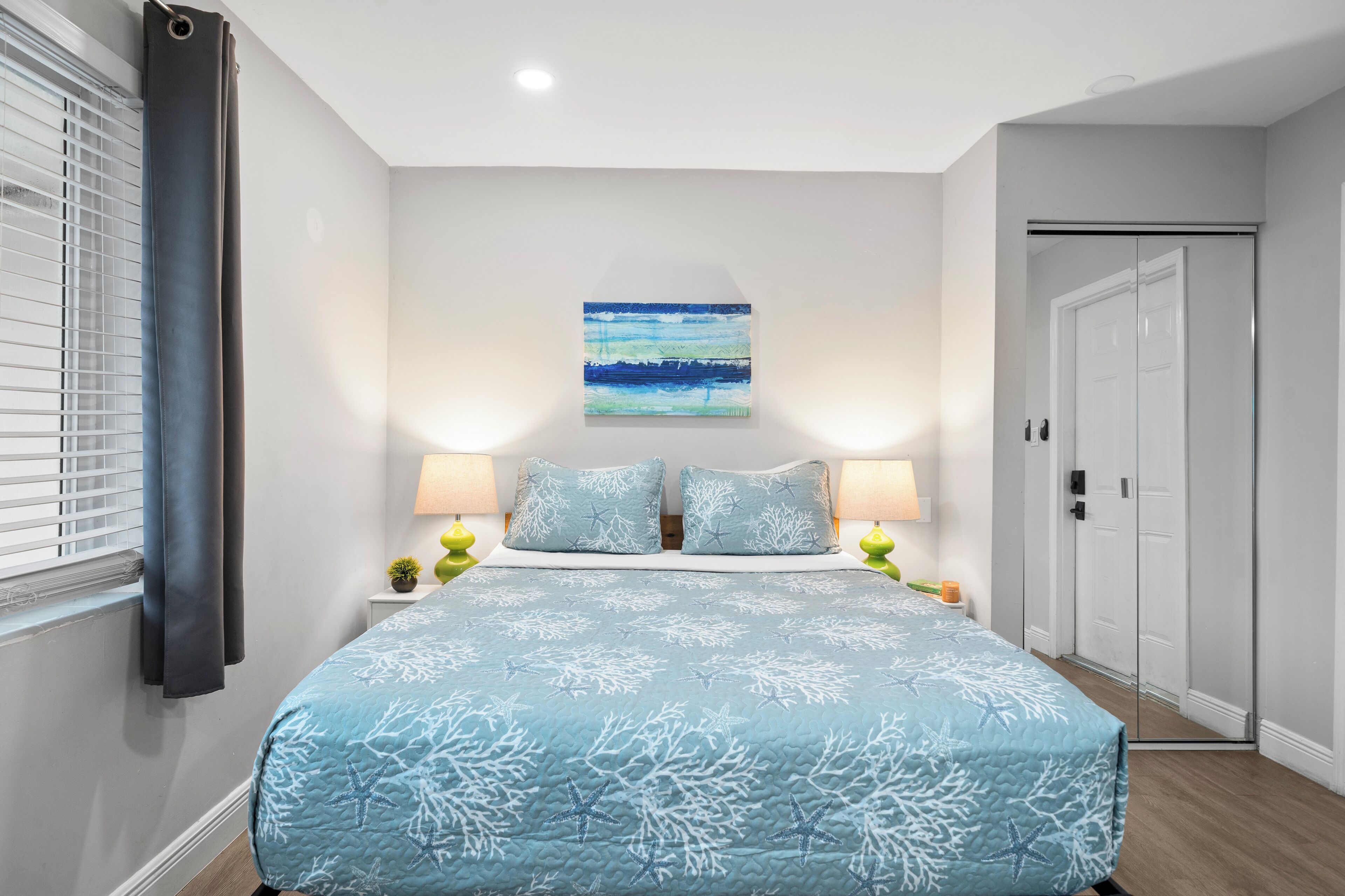 A bedroom with a bed, two bedside lamps, a nightstand, a window with curtains, a wall-mounted painting, and a door.