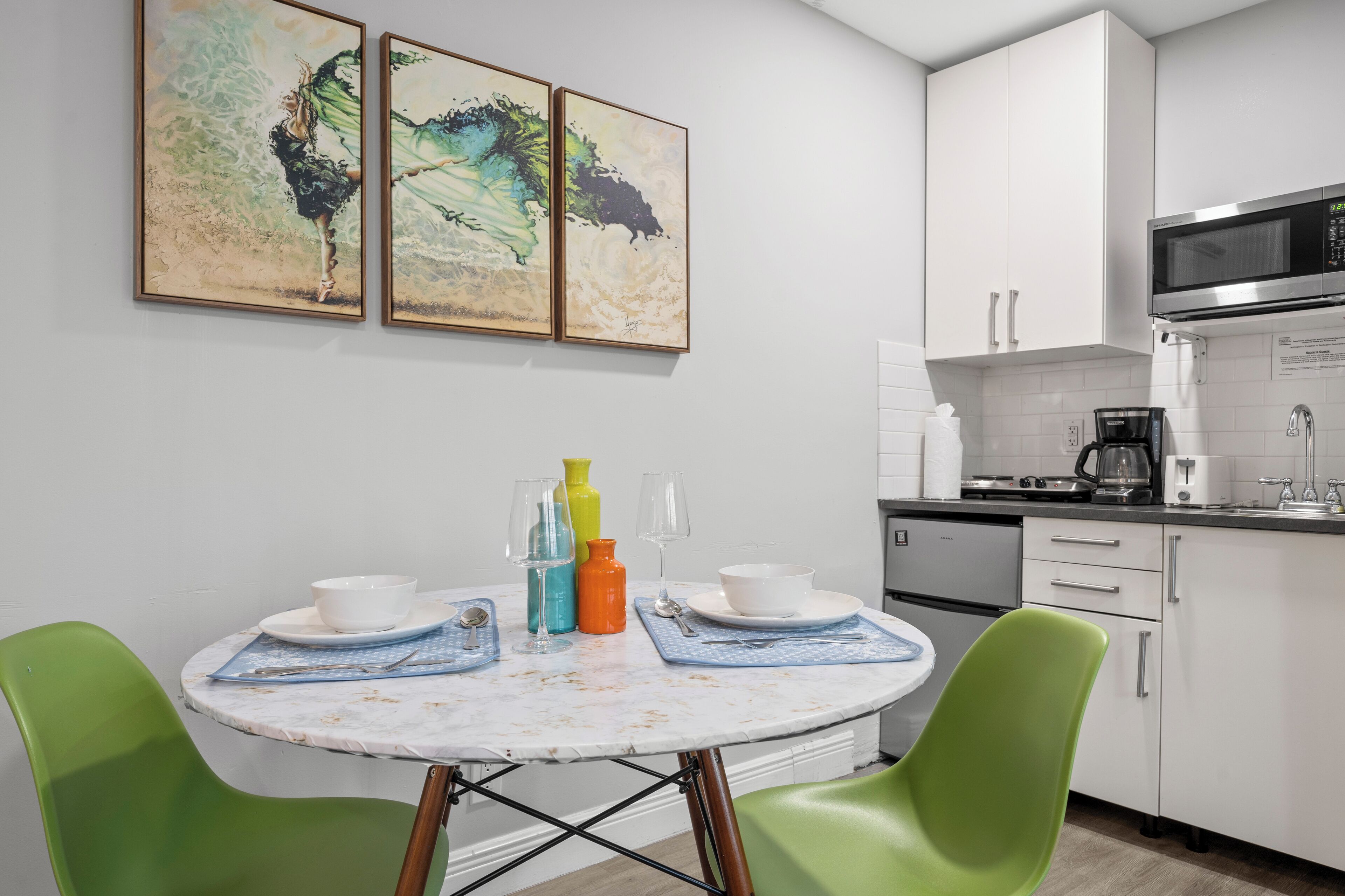 A compact kitchen with white cabinets, a microwave, and a coffee maker. There is a dining table with green chairs and a white tablecloth.