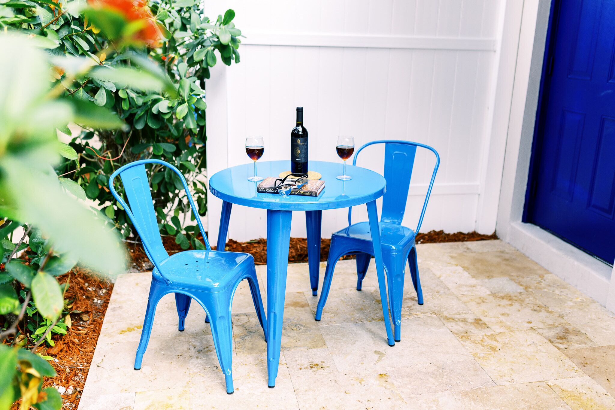Ein blauer Tisch mit zwei Stühlen, einer Flasche und zwei Gläsern Rotwein auf einer gefliesten Terrasse.