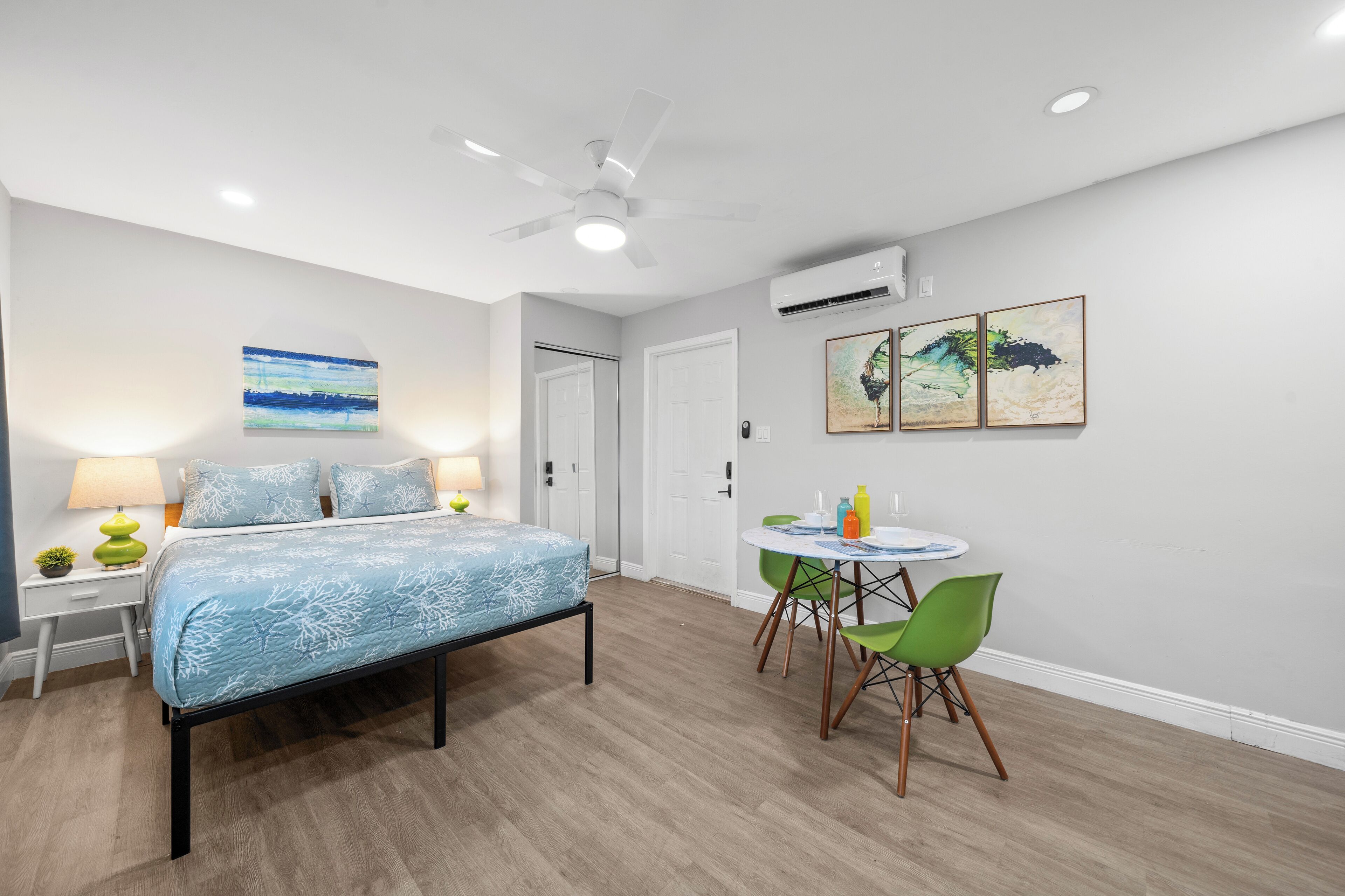 A bedroom with a bed, bedside table, lamp, and a small plant. There is a door and an air conditioning unit on the wall.