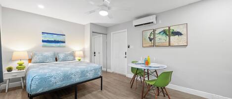 A bedroom with a bed, bedside table, lamp, and a small plant. There is a door and an air conditioning unit on the wall.