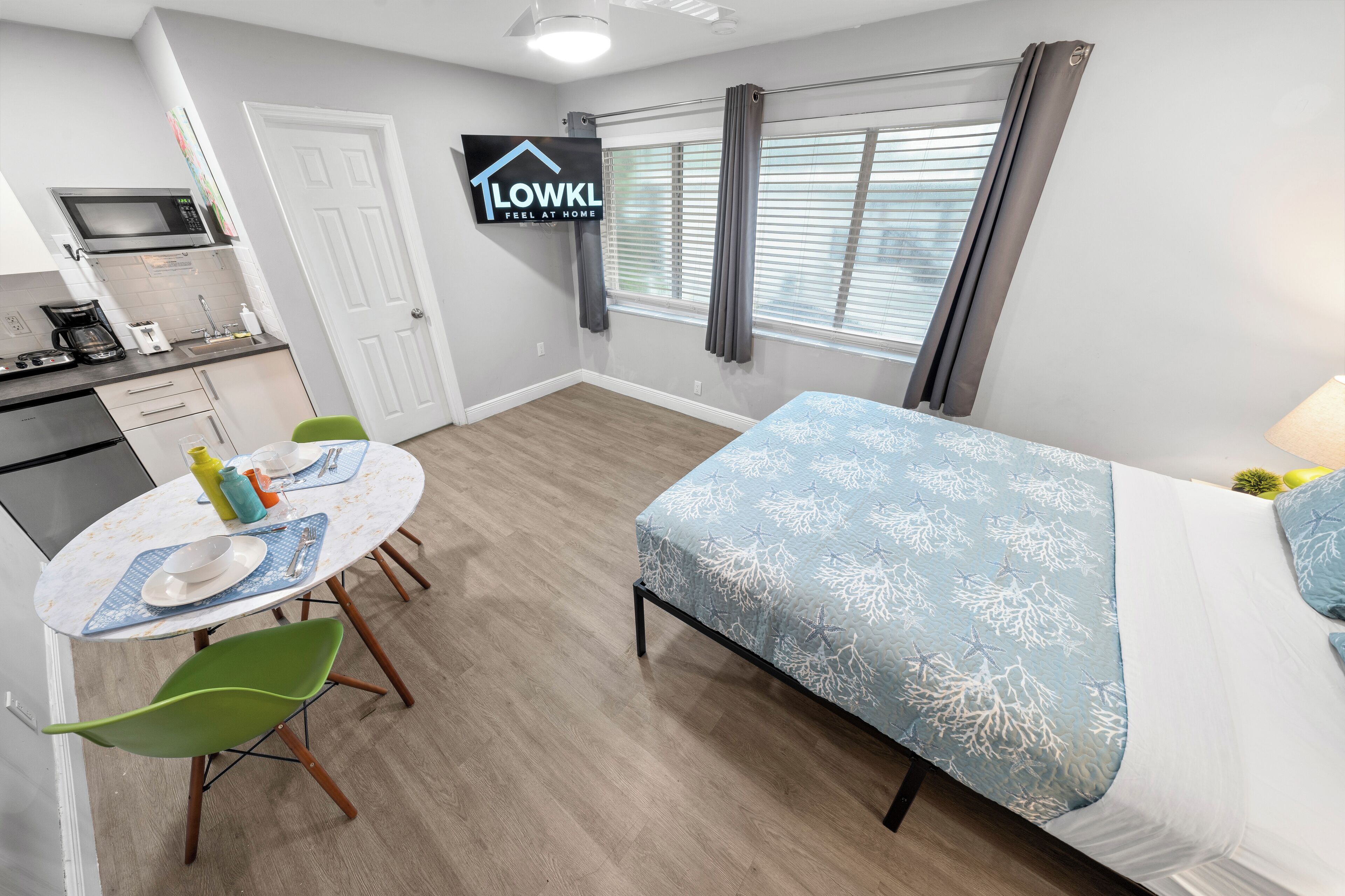 A compact hotel room with a bed, a dining table with chairs, a microwave, and a TV displaying a 'LOWK'L' sign.