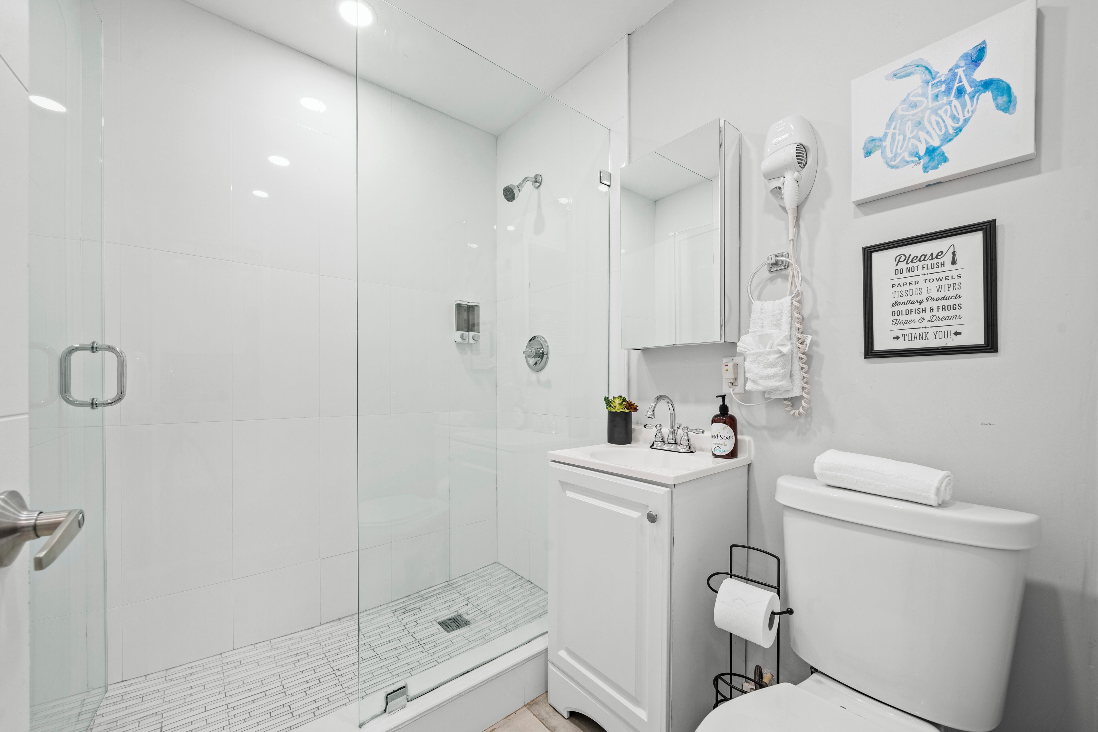 A modern bathroom with a glass-enclosed shower, a white sink with a mirror, and a toilet. There is a framed sign on the wall and a towel rack.