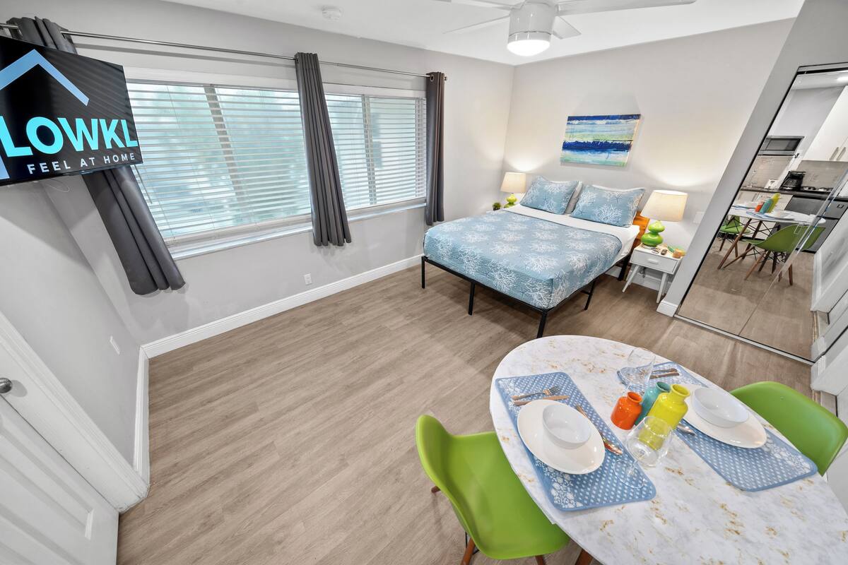 Ein modernes Hotelzimmer mit einem Bett, einer Essecke mit Tisch für zwei Personen, einer Küchenzeile und einem großen Fenster mit Vorhängen.
