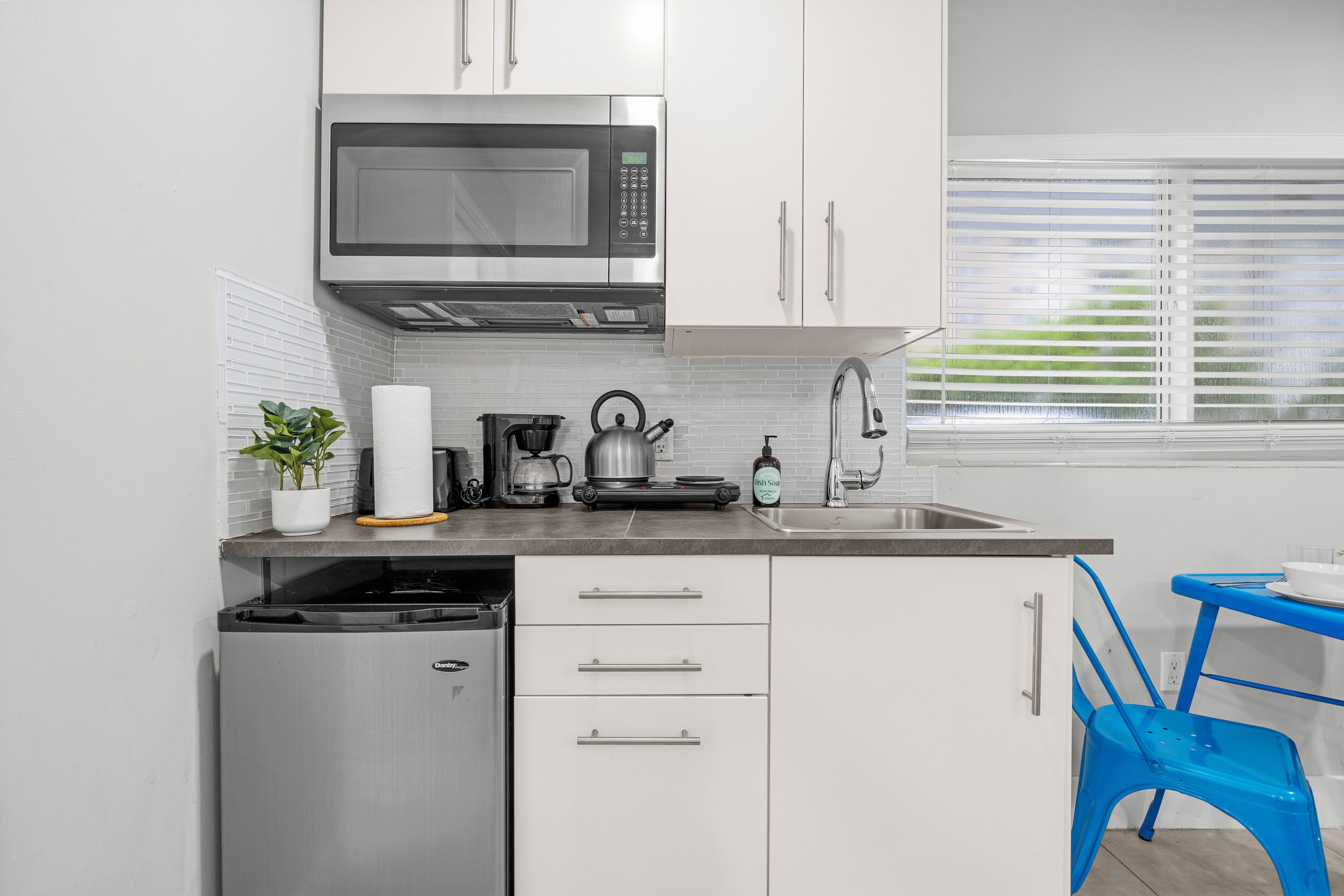A compact kitchen with a built-in microwave, a coffee maker, and a kettle on the counter. There is a small refrigerator and a blue chair.