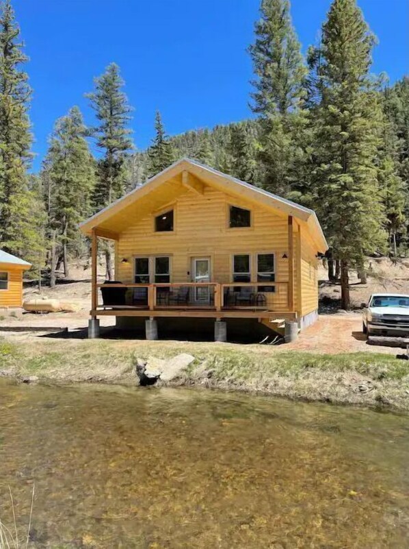 A wooden cabin with a balcony, situated by a clear stream, surrounded by tall pine trees.
