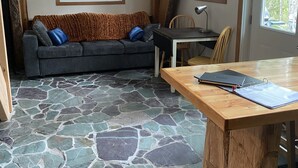 Una habitación acogedora con suelo de piedra, techo de madera, un sofá con cojines, una mesa con una computadora portátil y ventanas que dejan entrar luz natural.