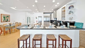 A modern kitchen with a central island, bar stools, and a dining area with a table and chairs. - Ultimate Paradise Retreat  (Oceanside)