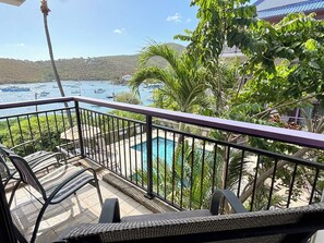 A balcony with a view of a swimming pool, surrounded by greenery and a hilly landscape.