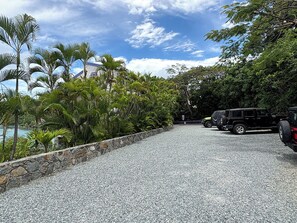 A gravel driveway with a stone wall, parked vehicles, and tropical vegetation. - This exceptional property offers the convenience of being just a short stroll from Cruz Bay, all while providing the comfort of full air conditioning. Don't miss out on this perfect blend of location and luxury! (St John)