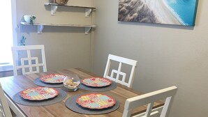 A dining area with a wooden table, white chairs, and a painting of a coastal landscape. - Lovely Beach View, 2 BR Condo (Corpus Christi)