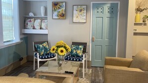 A living room with a ceiling fan, a coffee table with a vase of sunflowers, two sofas with patterned cushions, and a door with a light switch. - Lovely Beach View, 2 BR Condo (Corpus Christi)