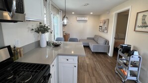 A compact kitchen with a stove, microwave, and white cabinetry. There's a dining area with a table and chairs, a gray sofa, and a coffee machine.