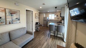 A modern living room with a grey sofa, a dining area with two chairs, a kitchen with a fridge and a TV mounted on the wall.
