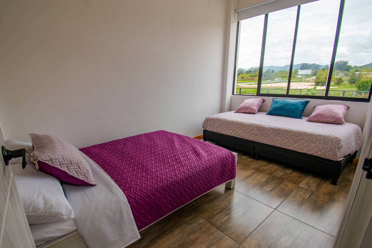 A bedroom with two beds, one with a pink bedspread and the other with a white bedspread. There is a large window providing a view of the outdoors.