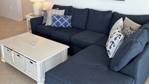 Un soggiorno con un divano angolare grigio scuro, un tavolino da caffè bianco e un quadro raffigurante una spiaggia.