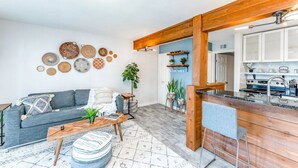 Un salon moderne avec un canapé, une table basse et des œuvres d’art décoratives au mur. On aperçoit à l’arrière-plan un coin cuisine avec des meubles en bois et un plan de travail.