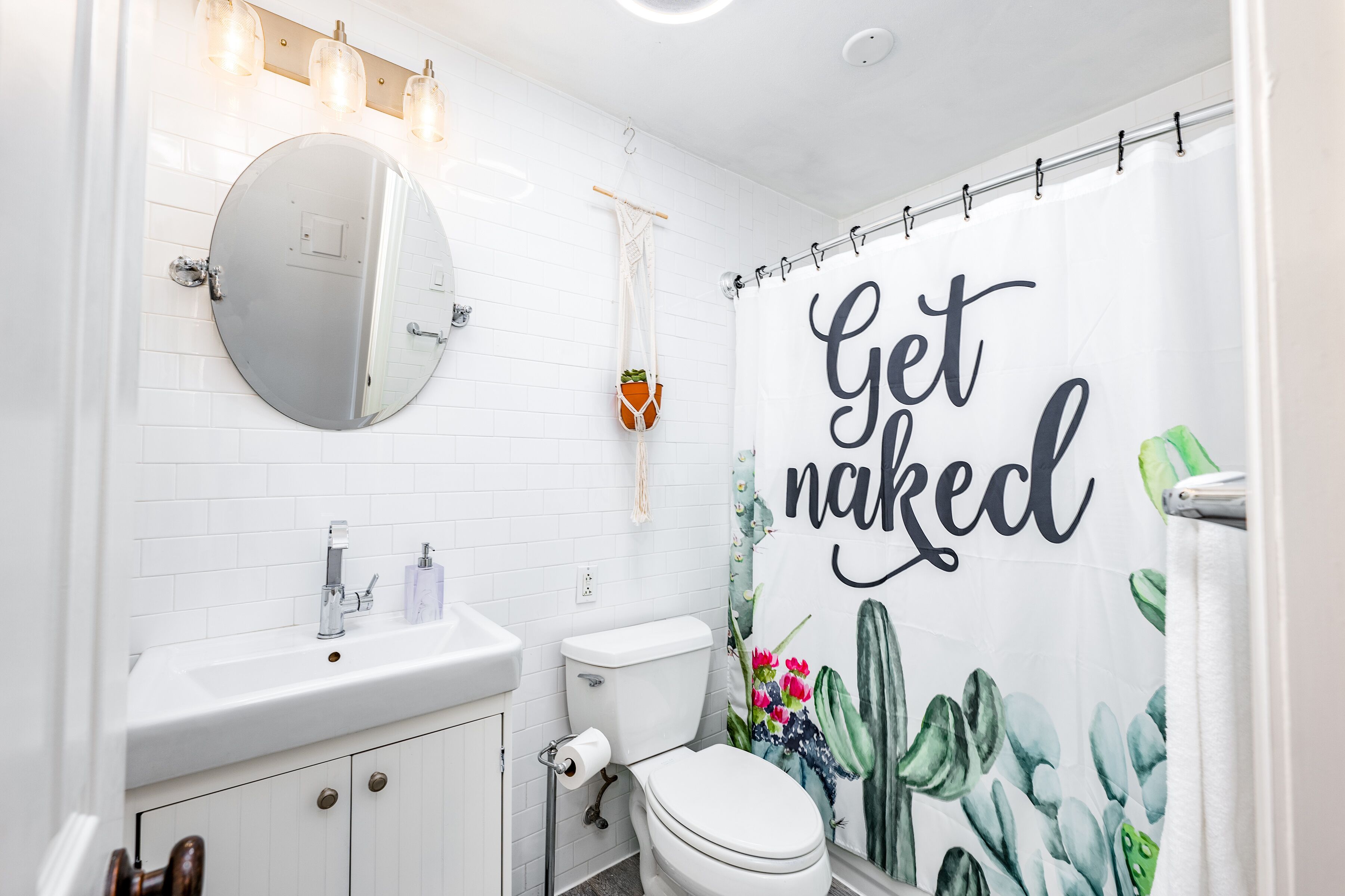 A bathroom with a white sink, mirror, and toilet, a shower curtain with a cactus design, and a hanging plant.