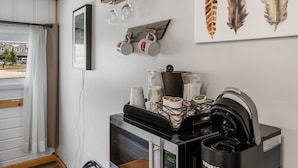 Une petite cuisine équipée d’un four à micro-ondes, d’une machine à café et d’une étagère où sont posées des tasses et une image de plumes.