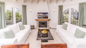 A living room with a fireplace, two white sofas, a marble coffee table with fruit, and large windows with curtains. - 2 Bedroom Villa in Landscaped Gardens (Javea / Xàbia)