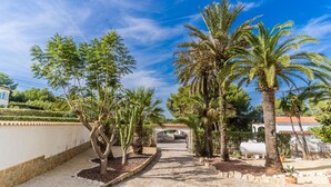 A pathway leading to a building with a tiled roof, surrounded by palm trees and a white wall. - 2 Bedroom Villa in Landscaped Gardens (Javea / Xàbia)
