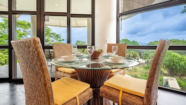 A dining area with a glass table surrounded by wicker chairs with yellow cushions, set against a backdrop of large windows overlooking a lush landscape. - EXCLUSIVE LA PERLA TAMARINDO CONDO 3BR LUXURY RETREAT, WALK TO BEACH AND DINING! (Tamarindo)
