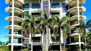 A modern multi-story building with balconies, a central atrium, and surrounding palm trees. - EXCLUSIVE LA PERLA TAMARINDO CONDO 3BR LUXURY RETREAT, WALK TO BEACH AND DINING! (Tamarindo)
