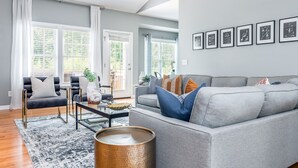 Een moderne woonkamer met een grijze bank, zwarte fauteuils en een salontafel. Er zijn grote ramen met witte gordijnen, een zwart-wit tapijt en lijstwerken aan de muur.