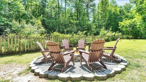 Een ronde vuurkuil met vier Adirondack-stoelen eromheen geplaatst, omringd door een stenen rand, gelegen in een gazon met een houten omheining en bomen op de achtergrond.