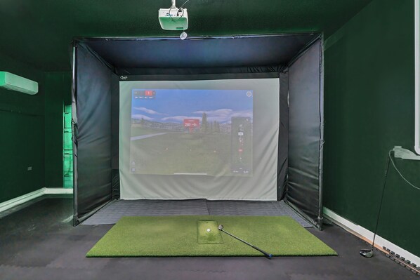 A golf training room with a screen displaying a golf course, a putting mat, and a golf club.