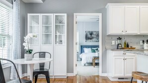 Een moderne keuken met witte kasten, een eethoek met een ronde tafel en stoelen, en een slaapkamer met een bed en een blauw hoofdeinde.