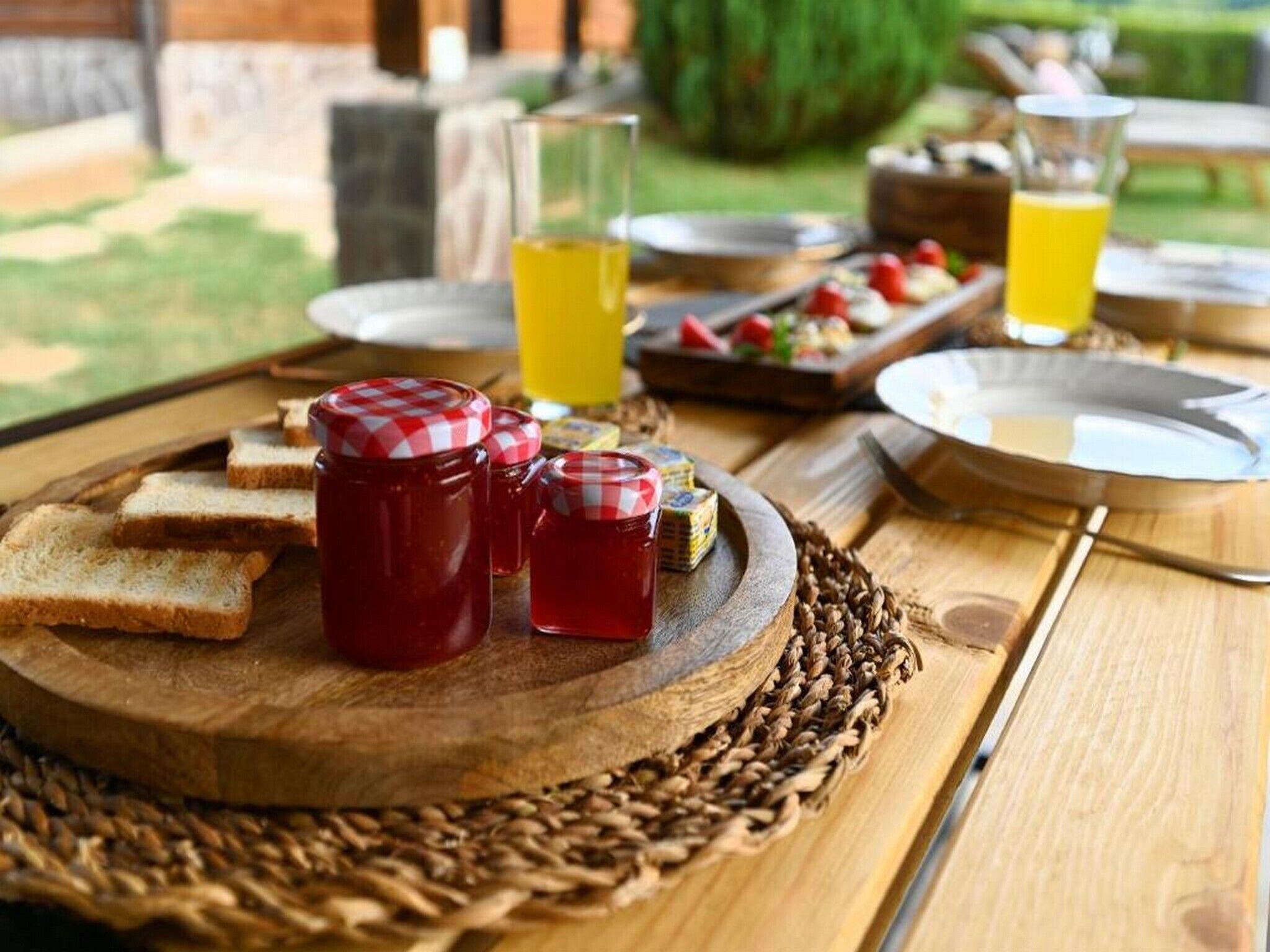 A wooden table with a woven placemat, containing jars of jam, slices of bread, and a glass of juice.