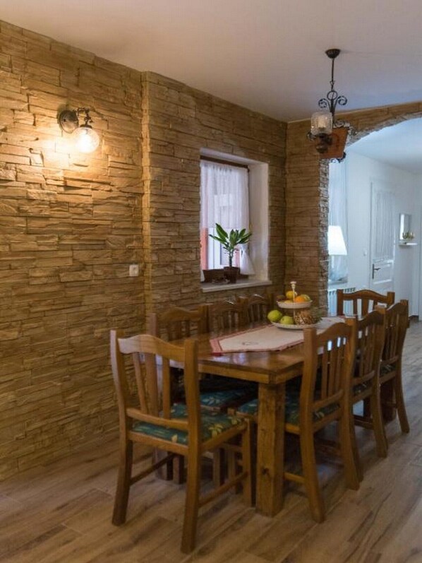 A dining area with a rustic stone wall, wooden table and chairs, a window with curtains, and a chandelier.