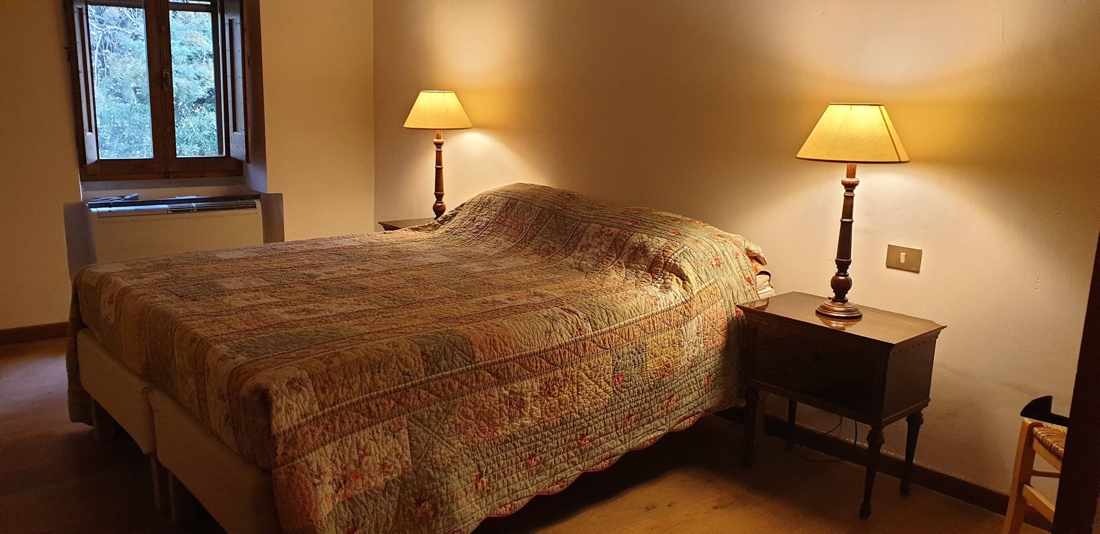 A bedroom with a large bed, two bedside lamps, a small table, and a window with a view of trees.