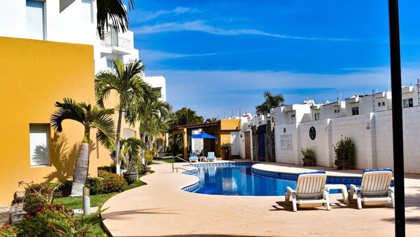 Una piscina con sillas de descanso, rodeada de palmeras y un edificio amarillo.