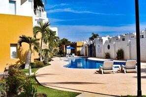 A swimming pool with lounge chairs, surrounded by palm trees and a yellow building. - Departamentos Arena (Mazatlán)