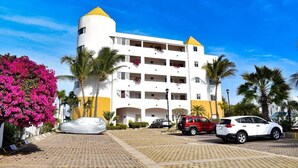 Un hotel de varios pisos con fachada blanca y detalles amarillos, con balcones adornados con plantas en macetas, un área de estacionamiento con varios autos y palmeras en primer plano.