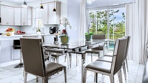 Eine moderne Küche mit weißen Schränken, ein Essbereich mit einem Glastisch und Metallstühlen sowie ein Blick auf einen Pool durch große Fenster.
